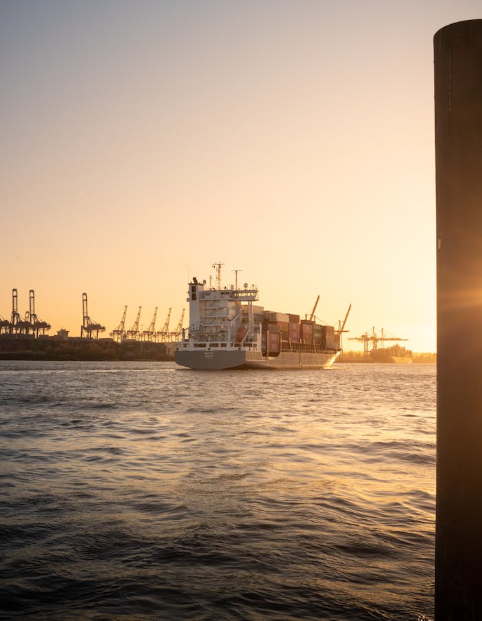 A container ship navigates through Hamburg harbor during a stunning sunset with cranes in the background.